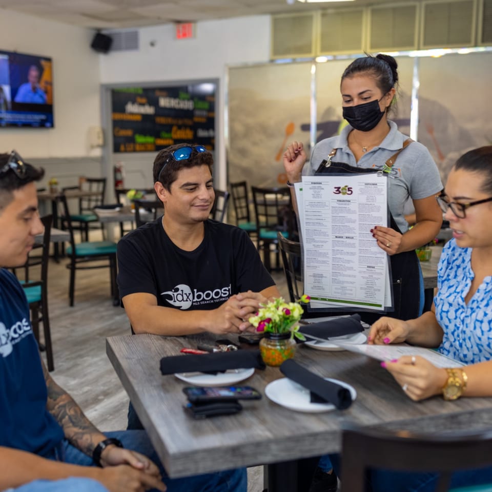 A server wearing a mask presents a menu to two men and a woman seated at a table.