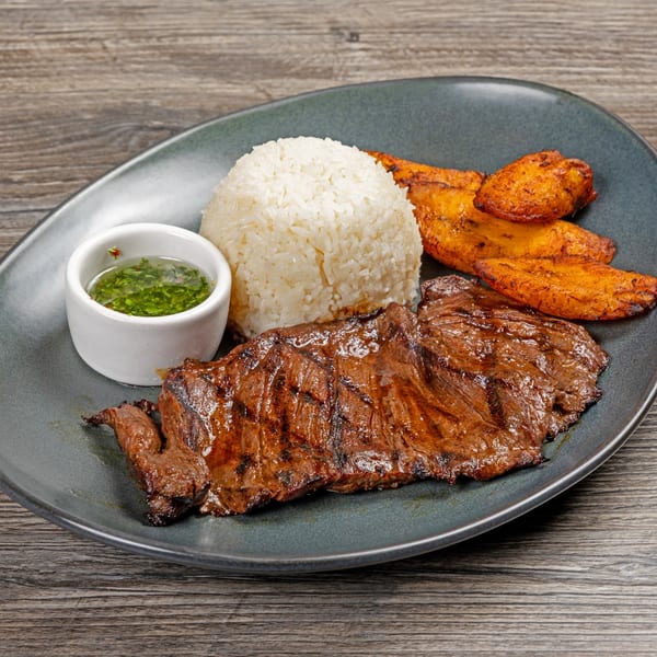 Peruvian grilled skirt steak with white rice, fried plantains, and green sauce on a gray plate.