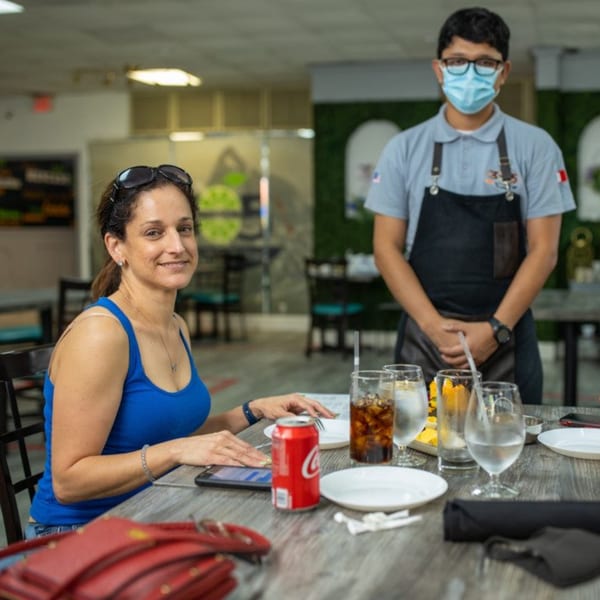 A smiling woman in a blue tank top sits at a table while a masked waiter stands behind her.