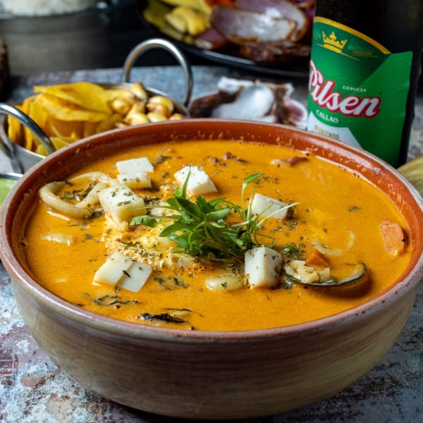 Peruvian seafood soup with cheese, herbs, and a Pilsen beer bottle in the background.