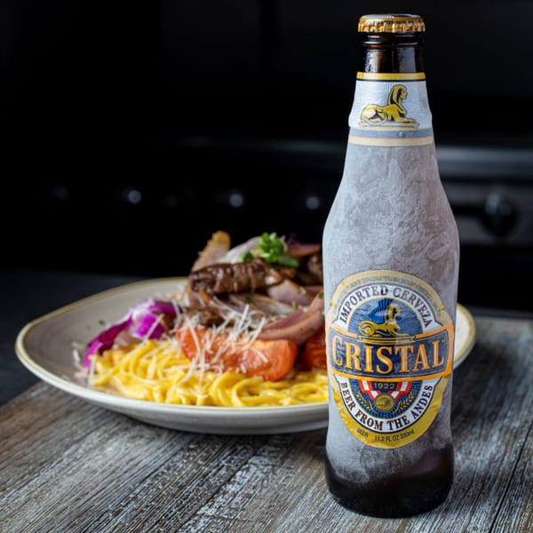 A frosty bottle of Cristal beer next to a plate of Peruvian lomo saltado with pasta.