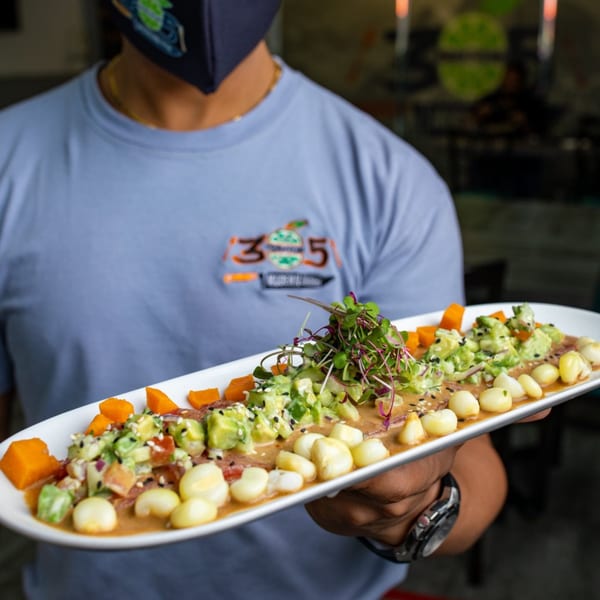 A server wearing a mask holds a long white plate of Peruvian ceviche with avocado, corn, and sweet potato.