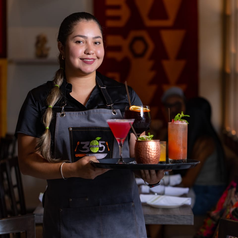 Smiling 305 Peruvian Modern Cuisine server holding a tray with four colorful cocktails.